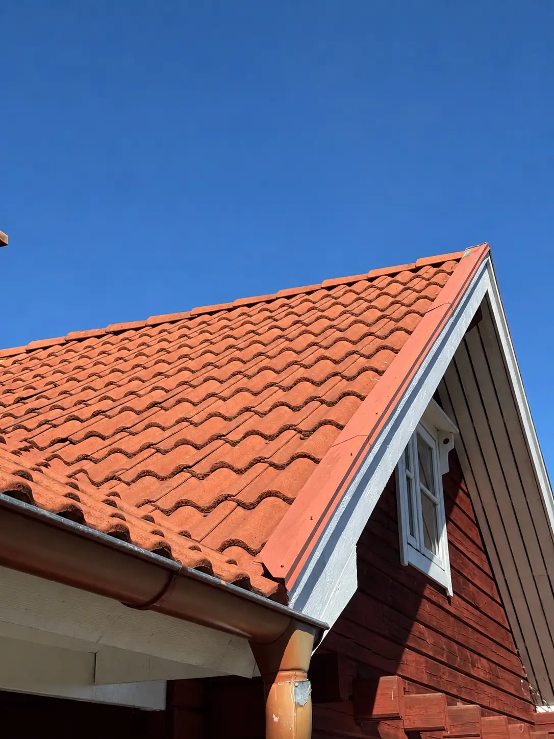 Rent rött tegeltak på rödmålad trävilla med vita fönsterfoder och ny nockplåt mot klarblå himmel